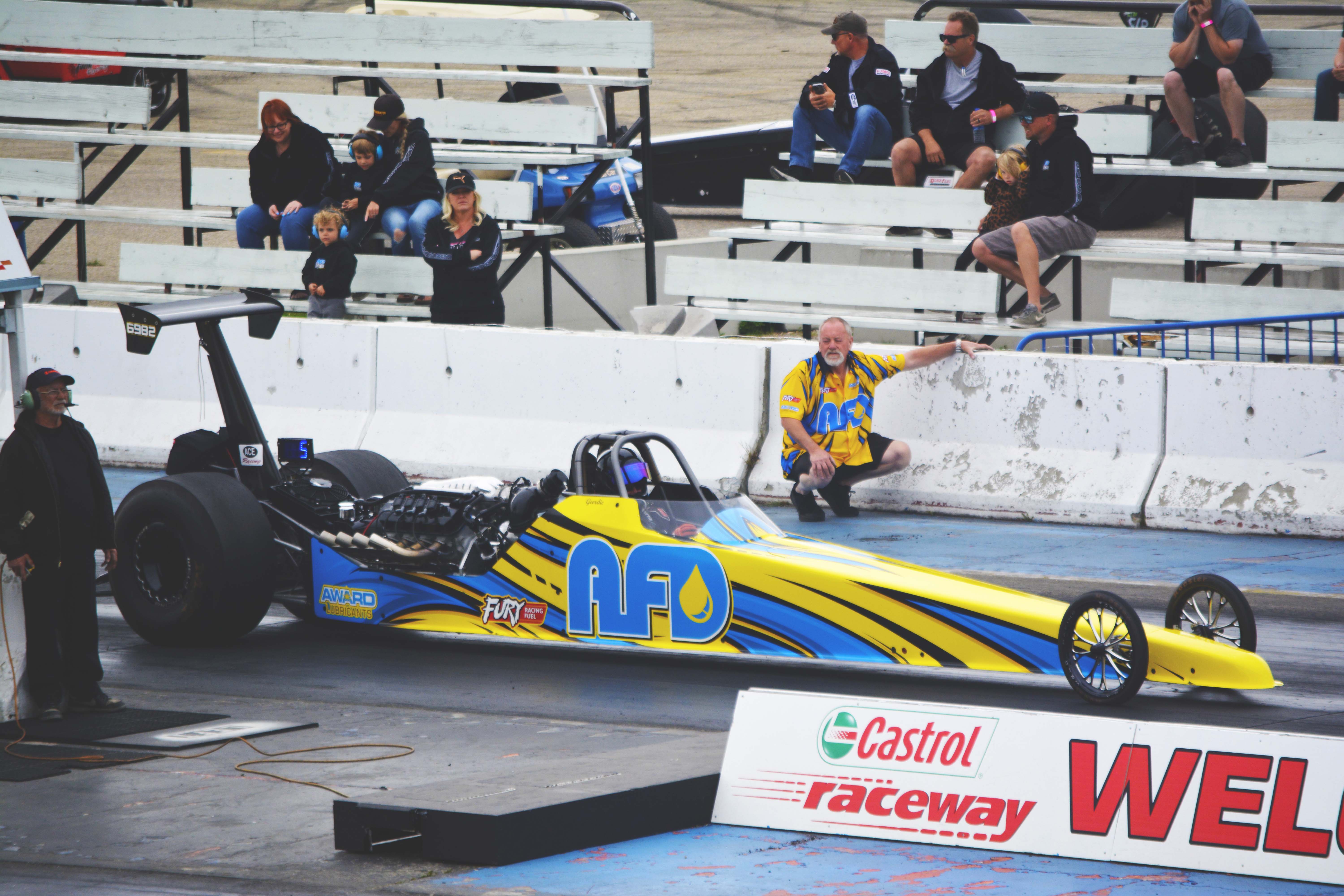 AFD Petroleum yellow dragster staged at Castrol Raceway with crew member and spectators