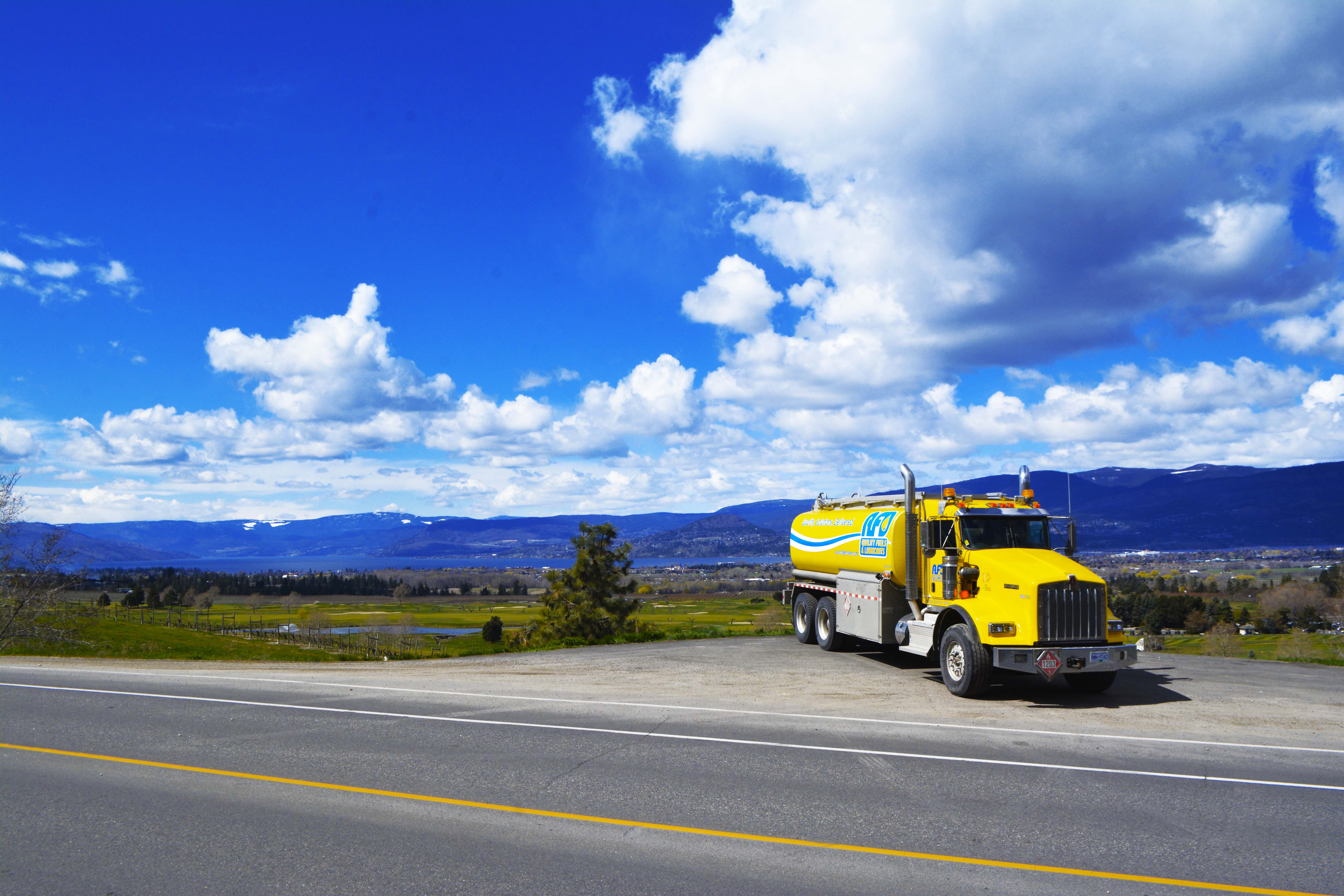 AFD Petroleum yellow fuel tanker truck parked on an overlook with a lake and snow-capped mountains behind it in Kelowna