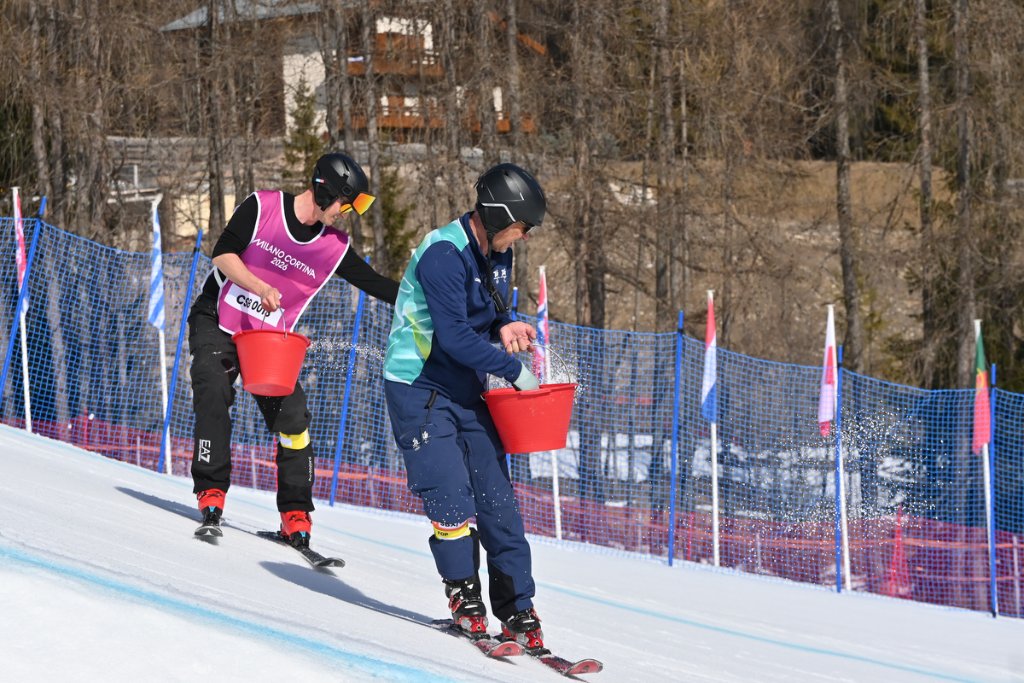 Course crew on skis pouring red buckets of water onto snowboard course at Milan Cortina 2026