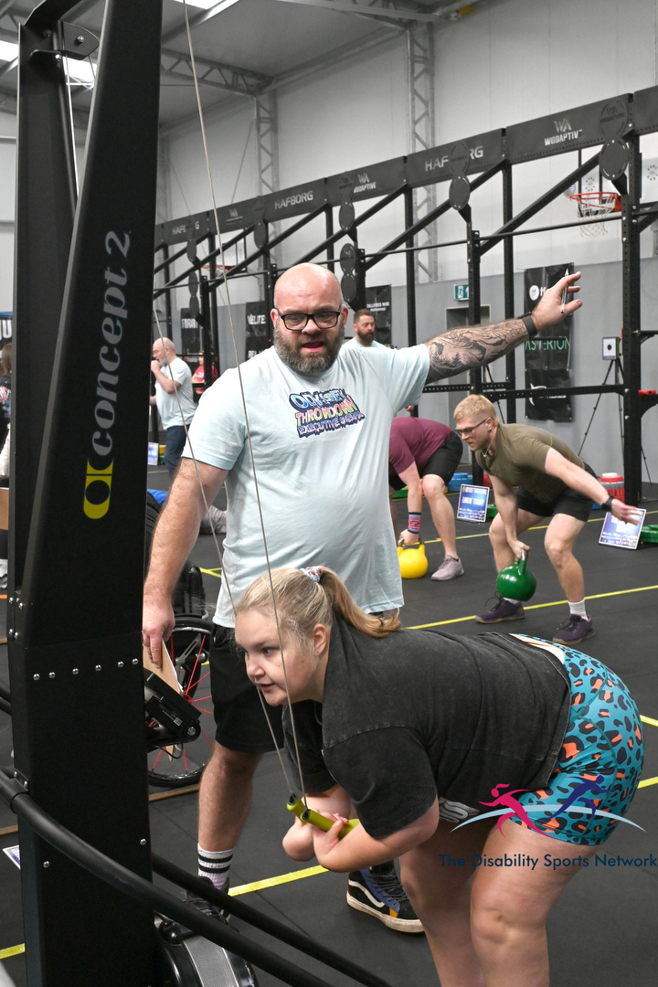 Athlete performing a cable exercise on a rowing machine while a coach spots her, at the Alt Movement adaptive fitness event