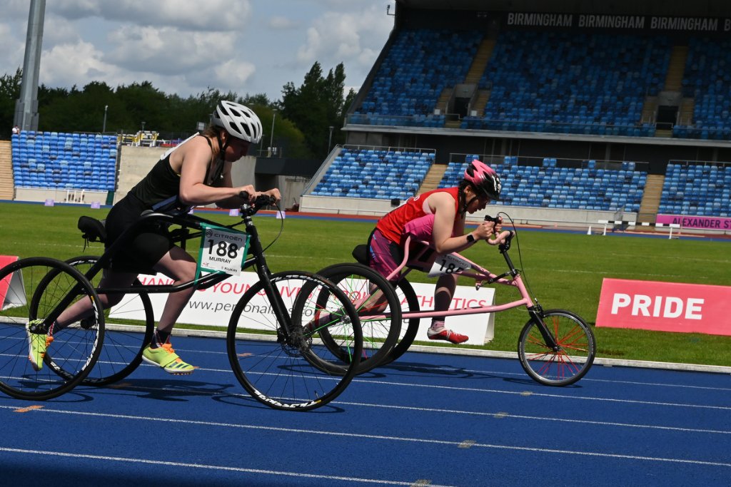Two para frame runners racing on the track with pride banner visible