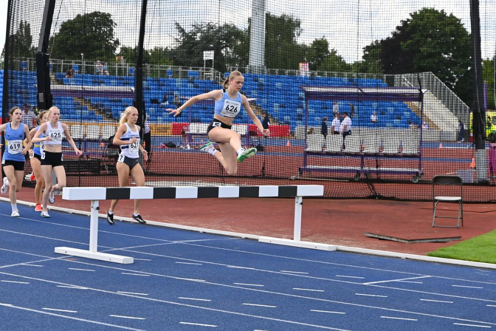 Female steeplechase athlete #626 clearing a barrier with competitors behind