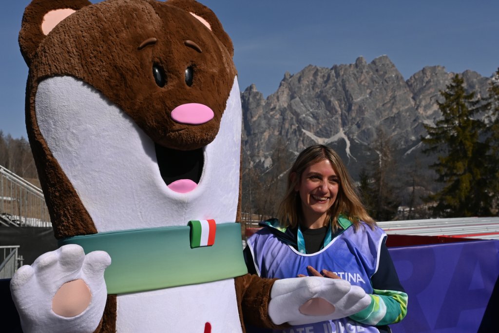 Race judge posing with Milan Cortina 2026 mascot in front of Dolomites mountains