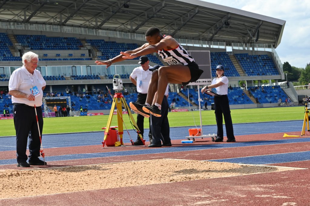 Male athlete in black and white at peak of triple jump with officials watching