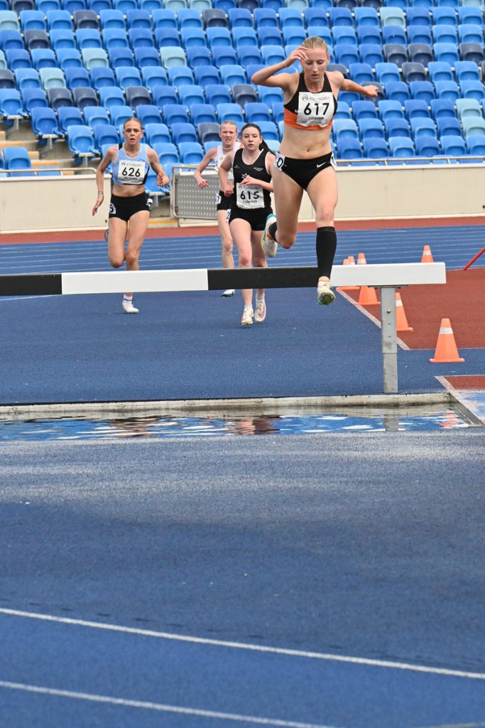 Female steeplechase athlete #617 leaping the water jump with competitors behind