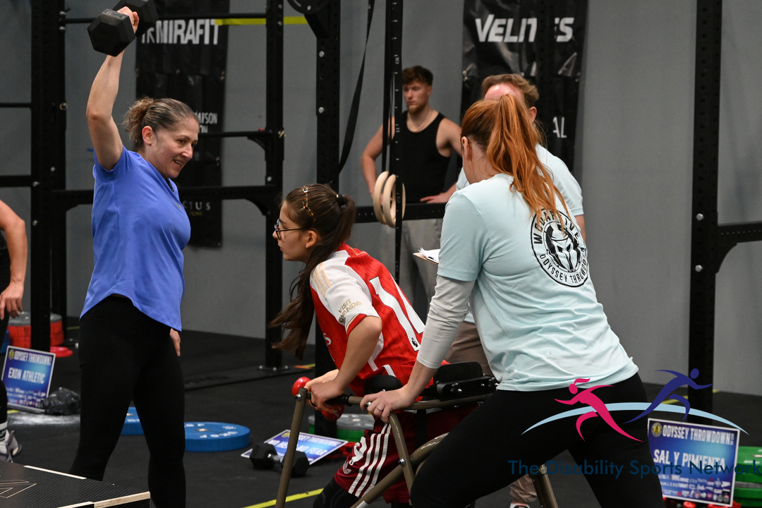 Athlete performing a one-arm dumbbell snatch while a coach guides another competitor at Alt Movement
