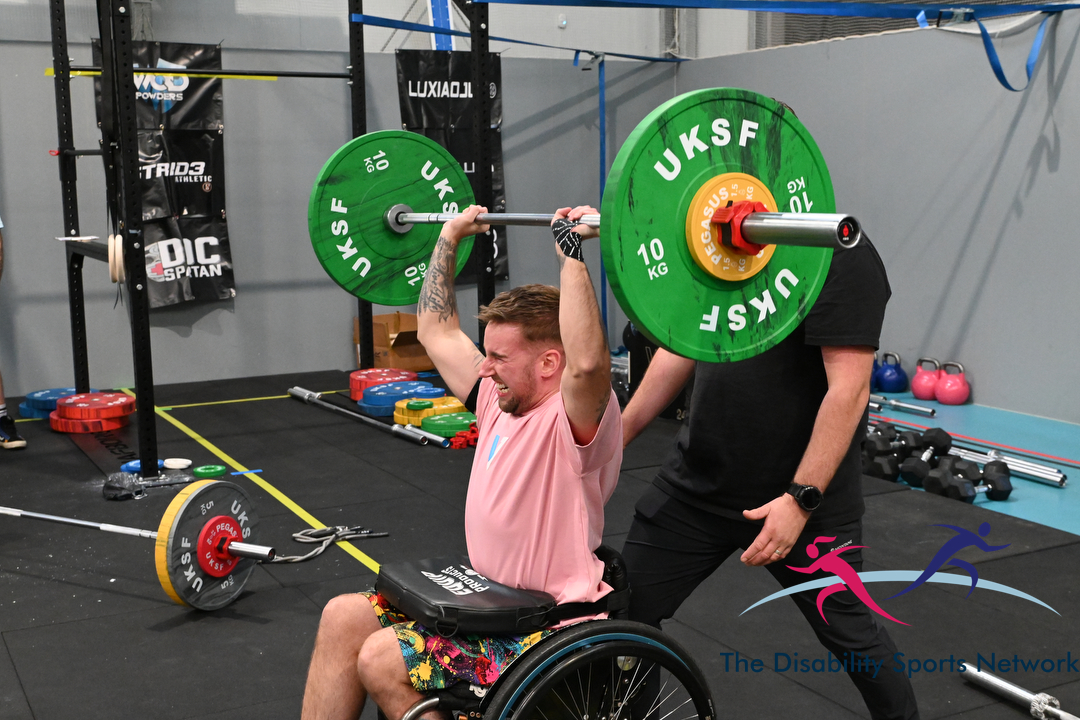 Wheelchair athlete performing an overhead barbell press with a spotter behind at Alt Movement