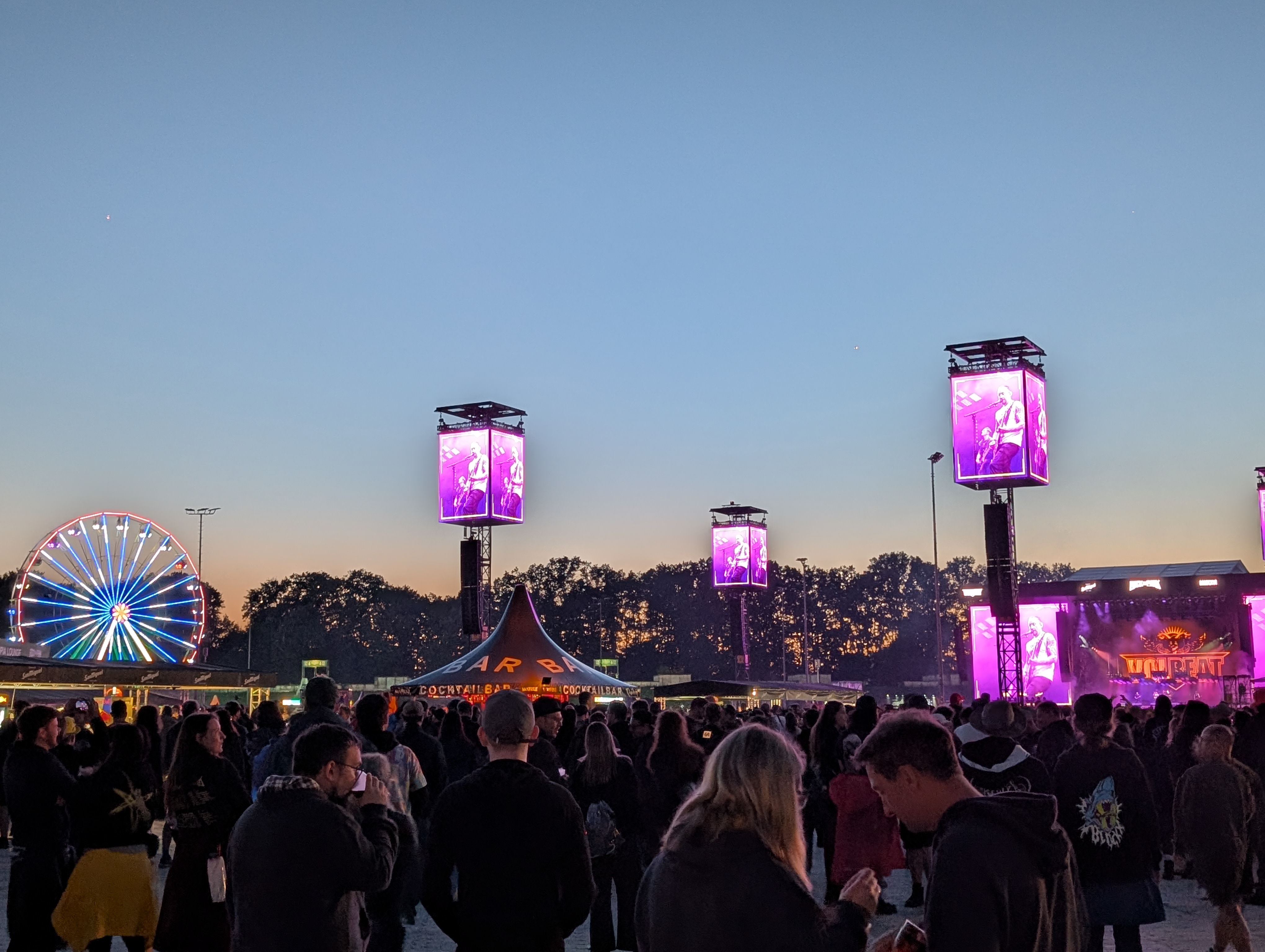 Festivalbühne im Abendlicht: große LED-Screens mit Live-Bildern, dichtes Publikum vor der Bühne und ein leuchtendes Riesenrad im Hintergrund