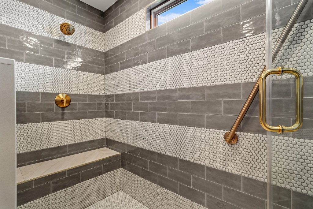 Inside view of Peacock Suite walk-in shower with grey subway tile, penny tile bands, brass showerhead, grab bar, and bench seat at Laurel Haven