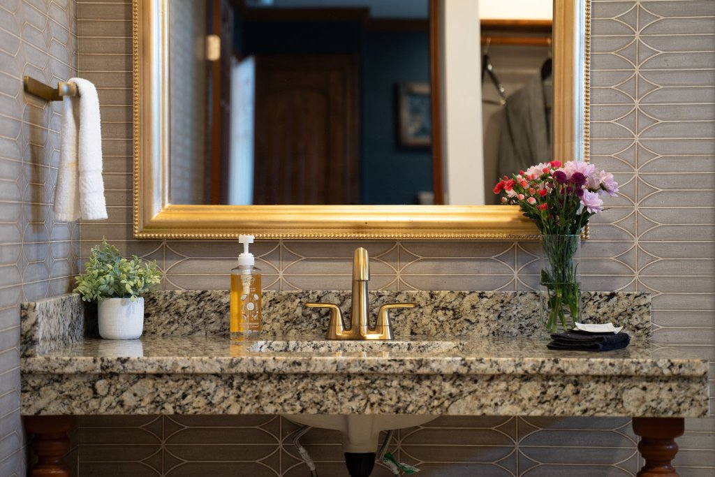 Peacock Suite bathroom vanity with granite countertop, brass faucet, gold ornate mirror, and geometric tile wall at Laurel Haven Estate