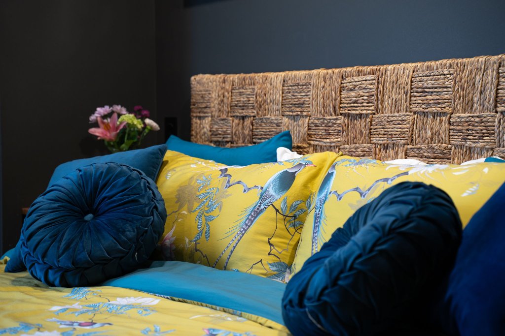 Close-up of Peacock Suite yellow peacock-print pillowcases with teal velvet round pillows and woven headboard at Laurel Haven Estate