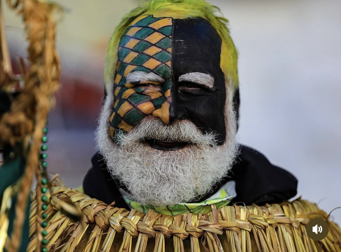 Close-up of a performer with green hair, harlequin face paint, white beard, and a woven straw collar