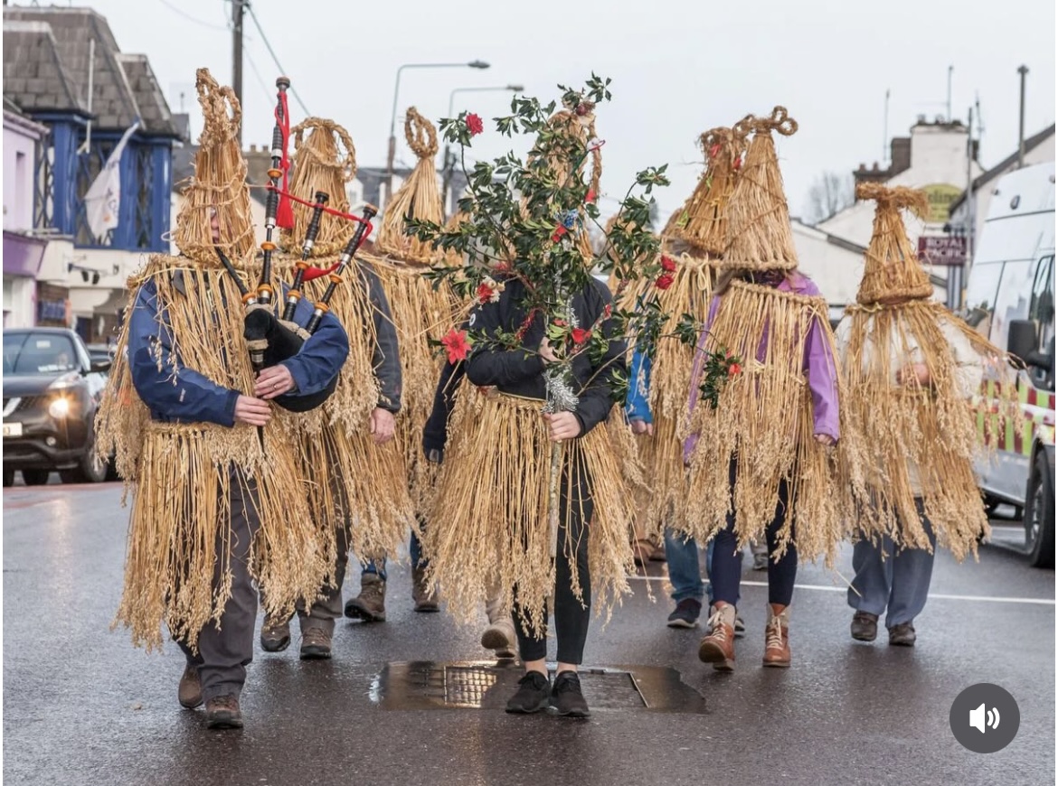 Straw-clad wren boys on a street, one playing bagpipes and another carrying a berried holly branch
