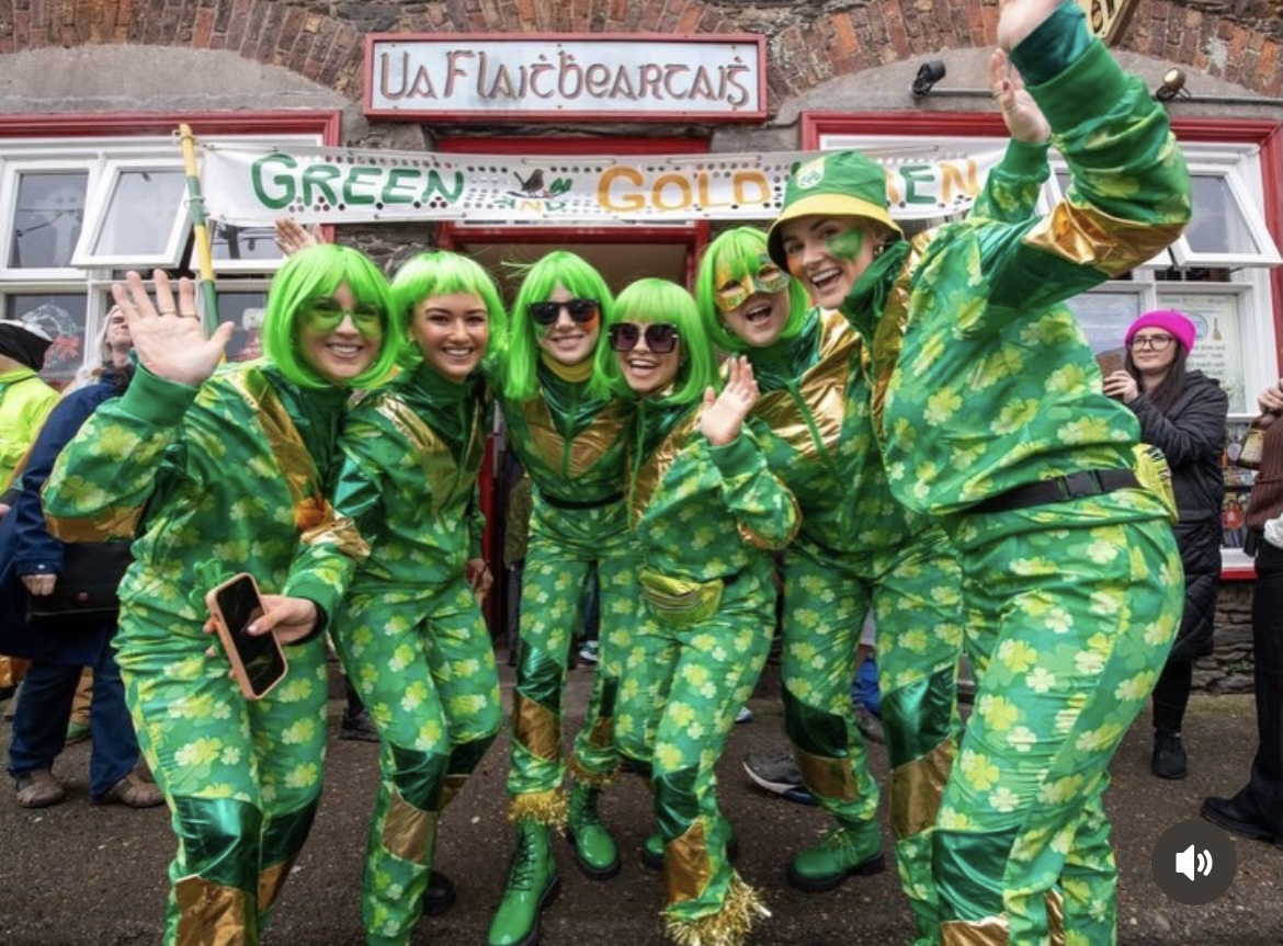 Six women in matching shamrock-print green tracksuits, lime wigs, and gold trim outside a GAA club