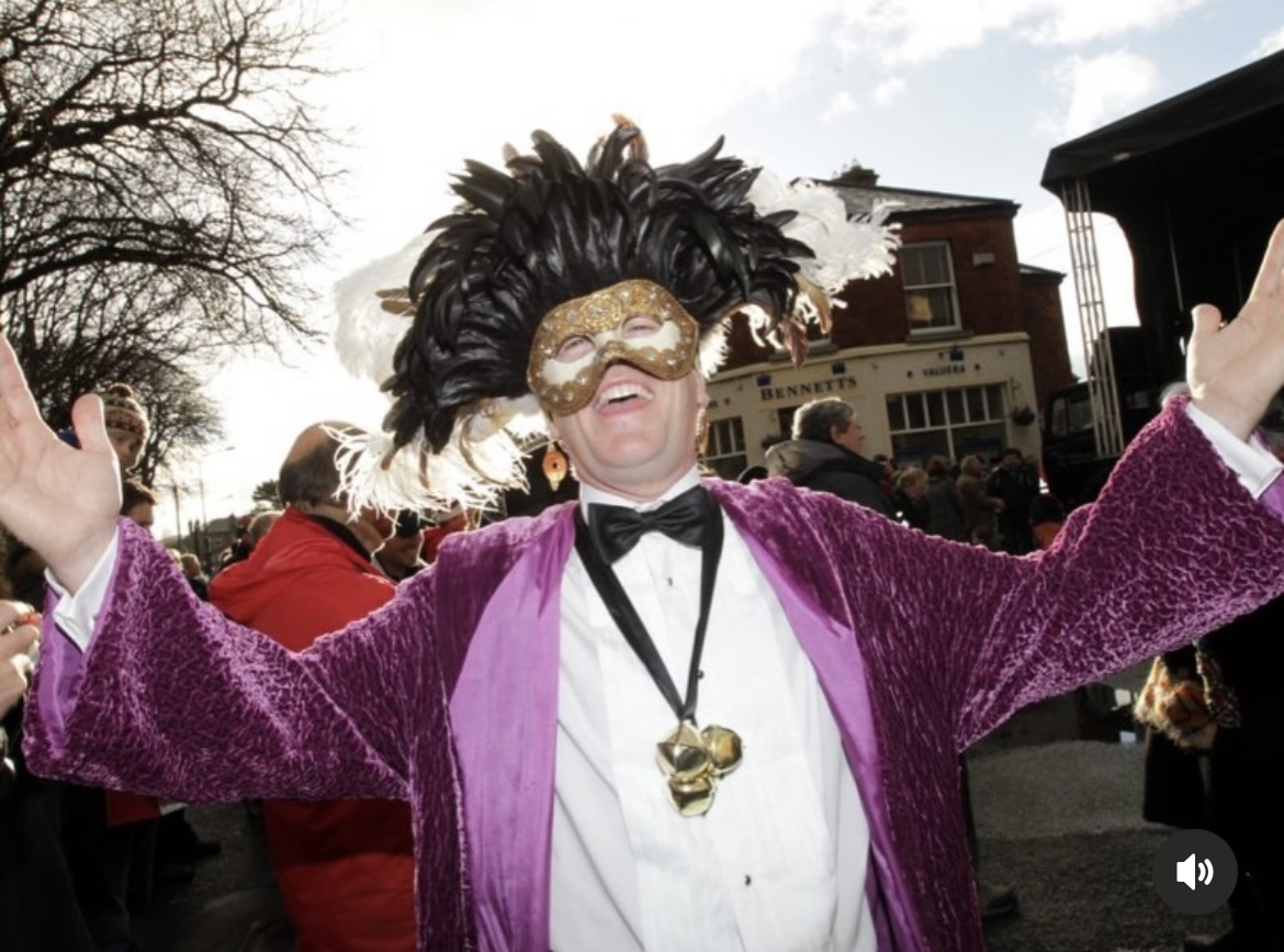 A performer in a purple velvet jacket, feathered headdress, gold mask, and brass sleigh bells