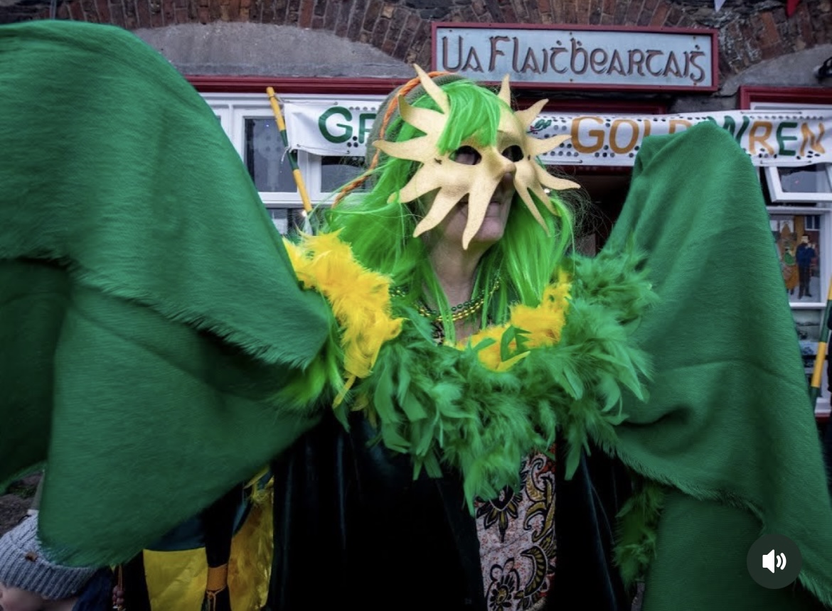 A performer in a sunburst mask, lime-green wig, feather boa, and wide green cape