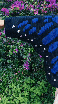Vídeo curto em loop mostrando o caimento do Cardigan Borboleta com asas em azul sobre fundo preto, cercado por vegetação e flores roxas