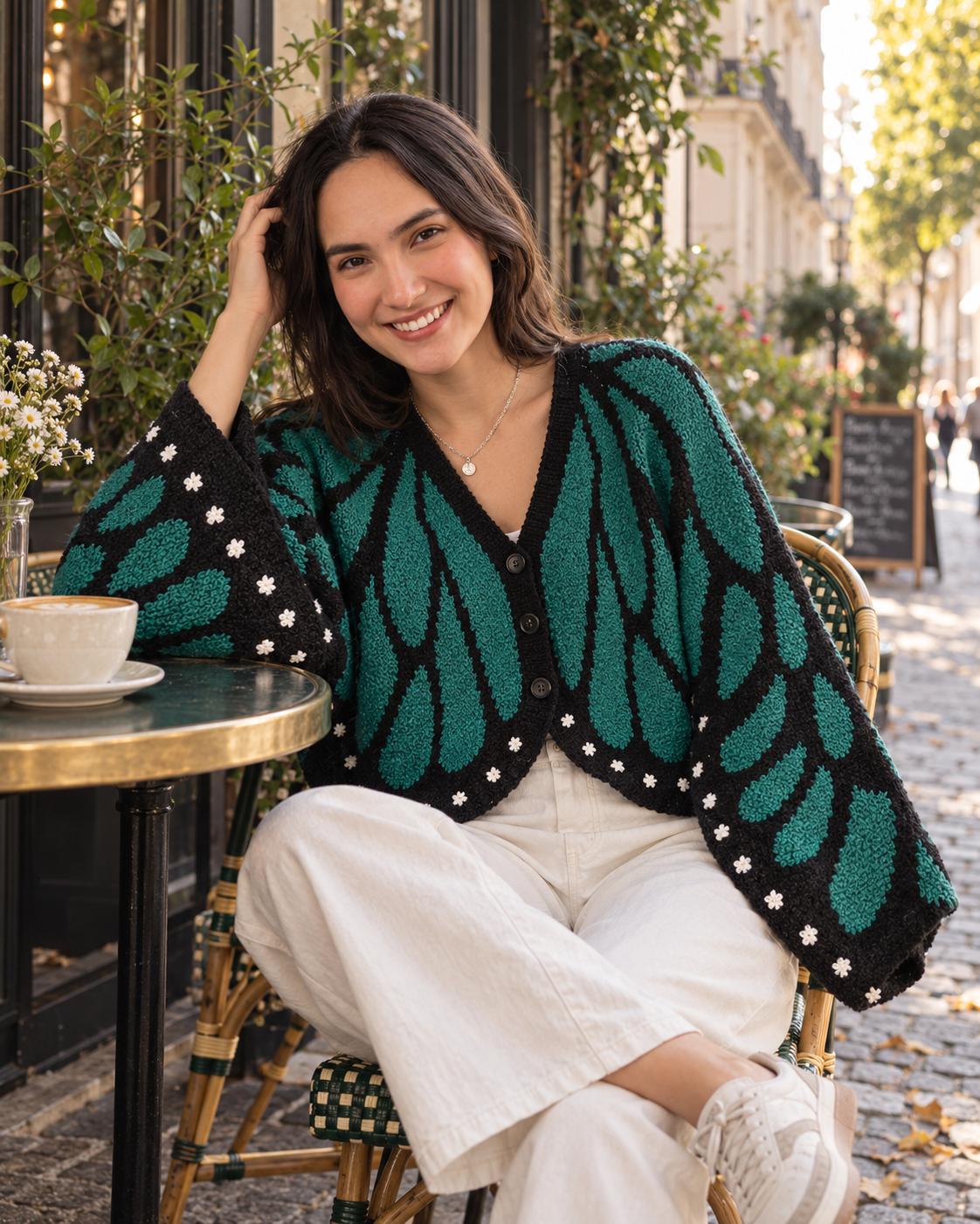 Mulher sorrindo usando o Cardigan Borboleta de crochê nas cores verde e preto, sentada em um café ao ar livre