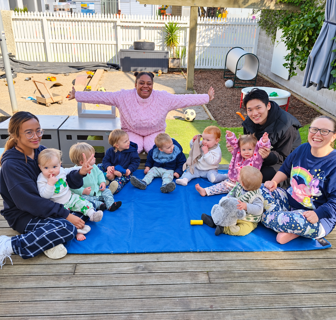 Teaching team and toddlers sitting together on a mat in the outdoor play area