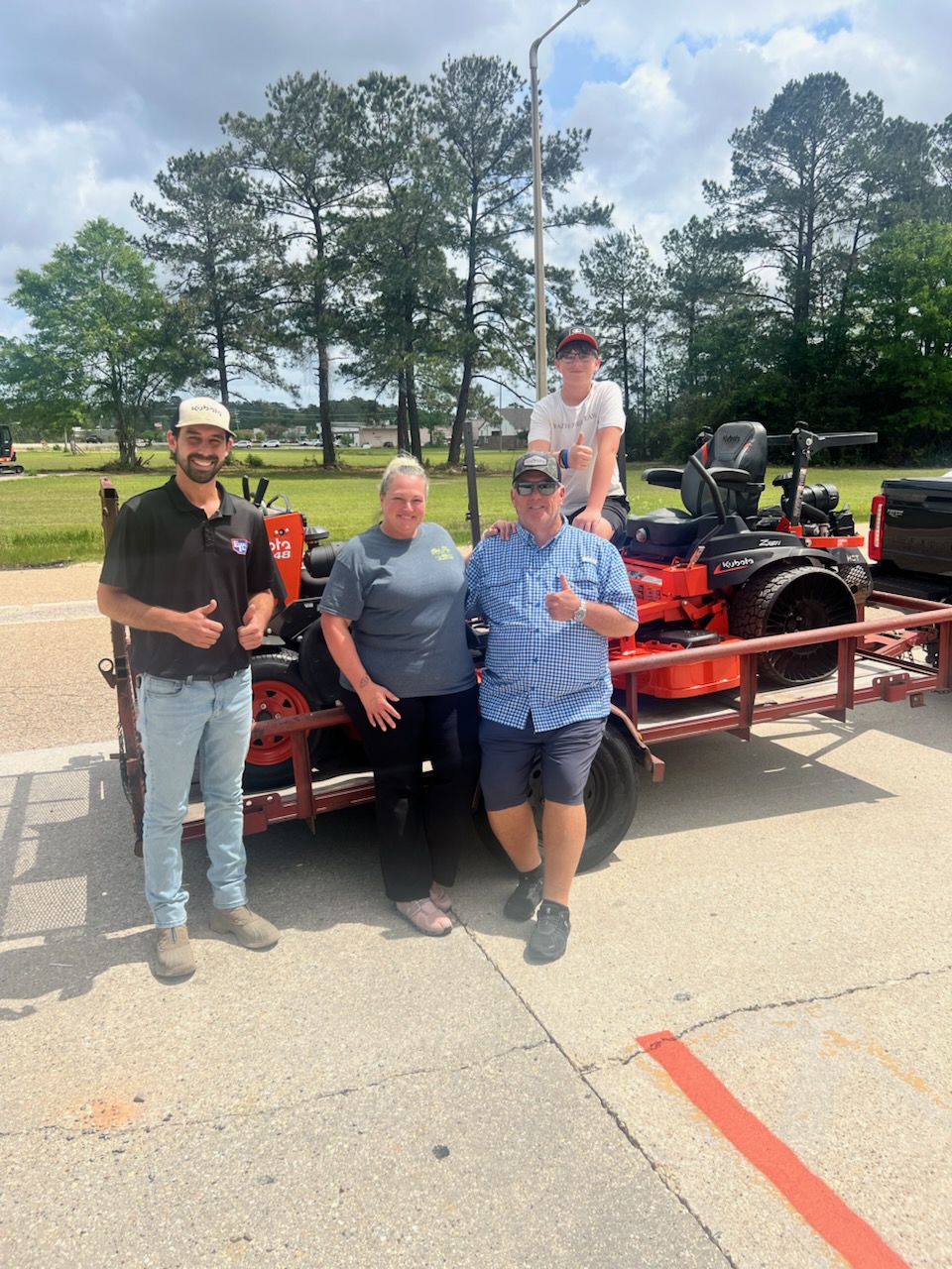Smiling customers giving thumbs-up in front of a Kubota zero-turn mower loaded on a trailer