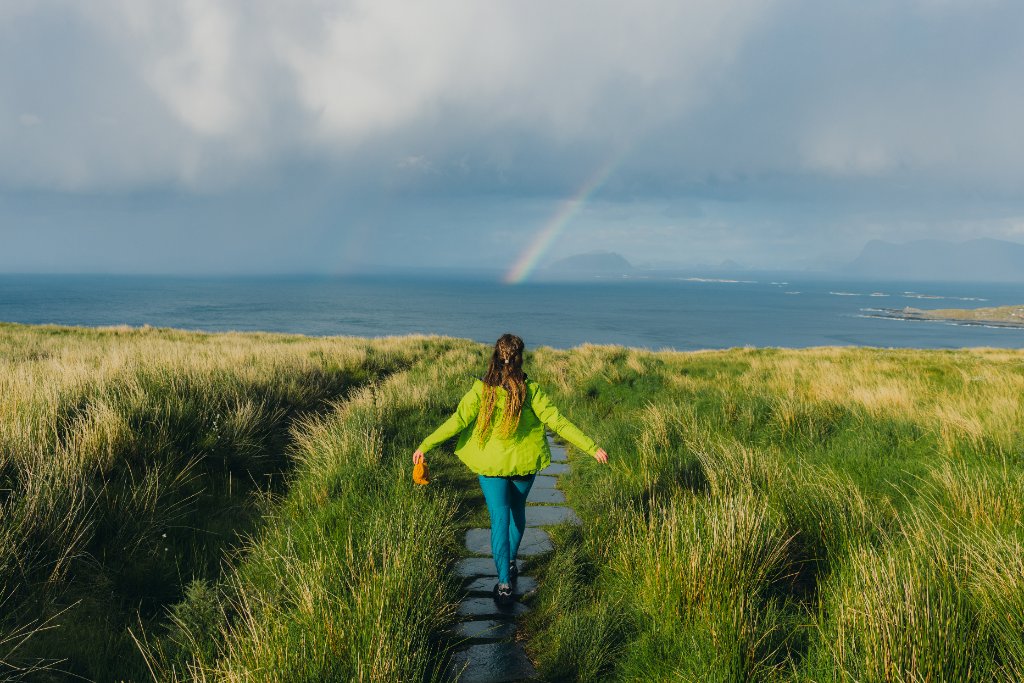 Woman walking a path through grass toward a rainbow over the ocean