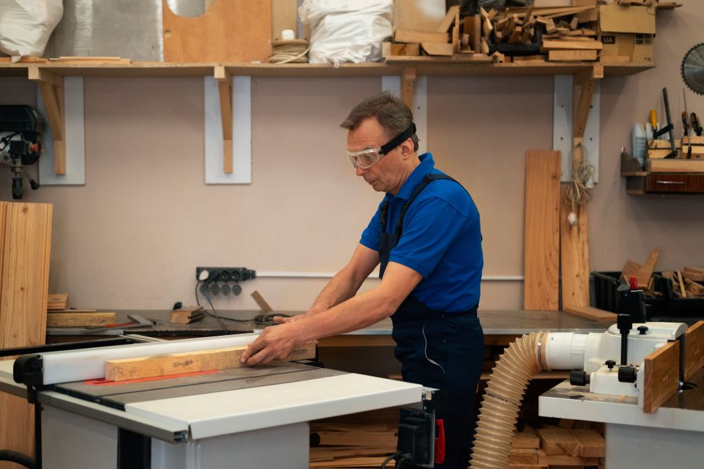 Craftsman in safety goggles guiding a wooden board through a table saw in a woodworking workshop