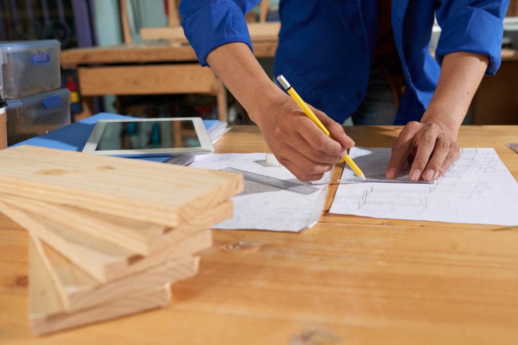 Craftsman in a blue shirt sketching technical plans with a pencil and ruler on a wooden workbench