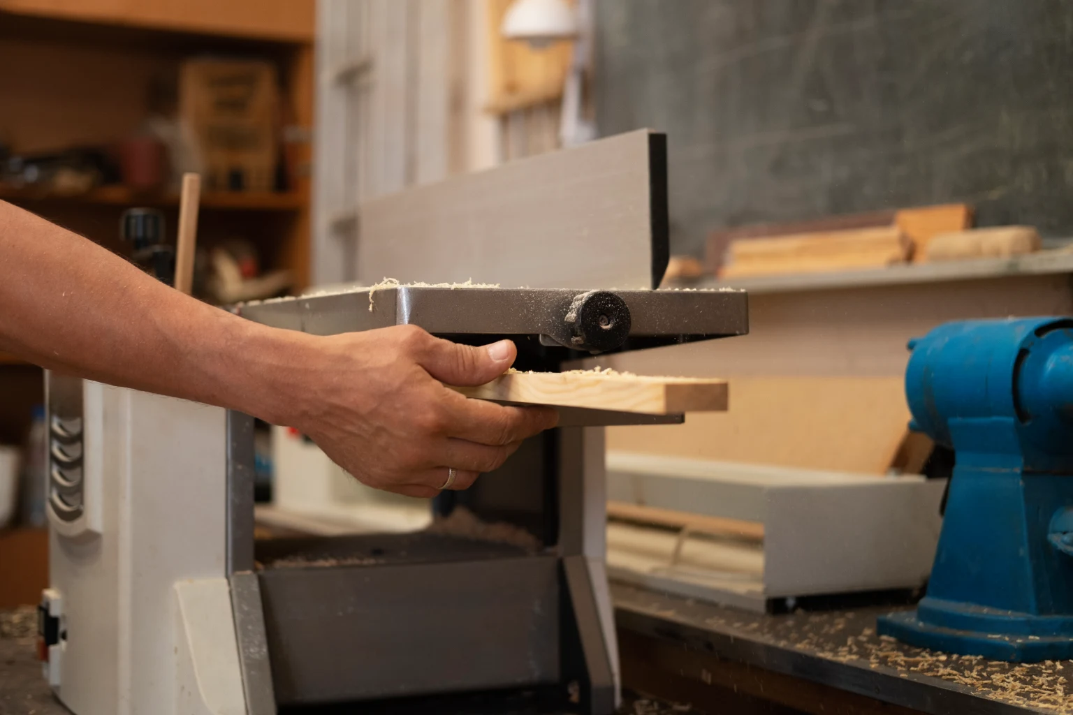 Person's hand feeding a piece of light-colored wood into an industrial wood planer in a workshop
