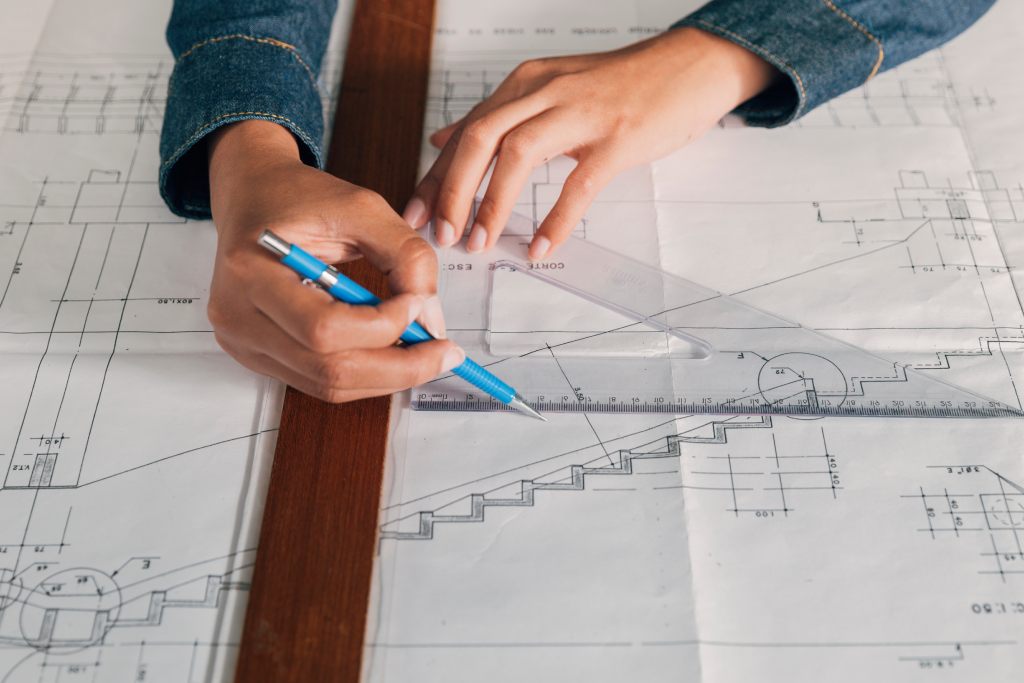 Hands drafting architectural plans with a blue mechanical pencil and transparent triangle ruler on white blueprints