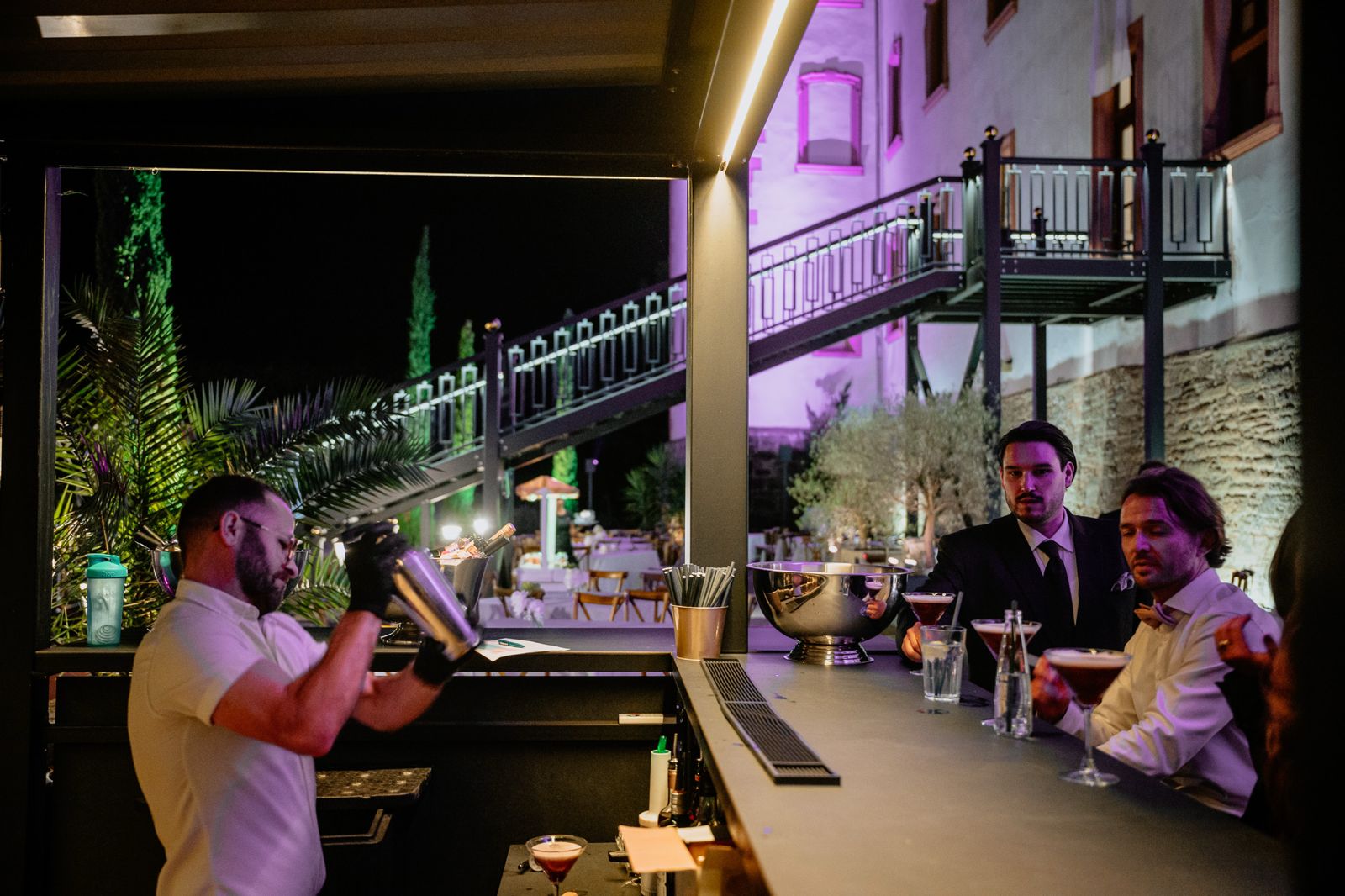 Outdoor-Bar mit angestrahlten Zypressen und Gästen bei Nacht