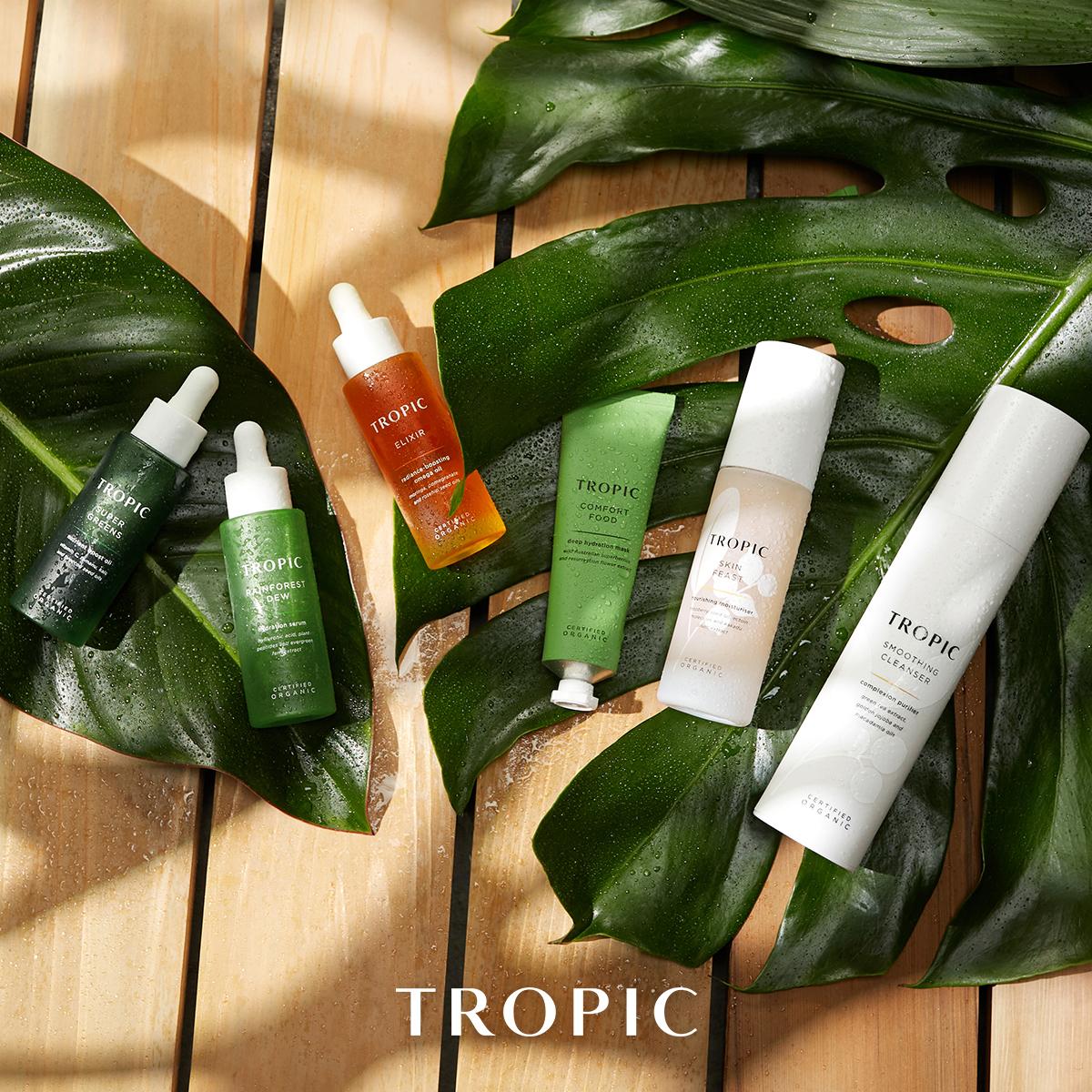 Flat lay of six Tropic skincare products, including Super Greens, Rainforest Dew, Elixir, Comfort Food, Skin Feast and Smoothing Cleanser, arranged on monstera leaves
