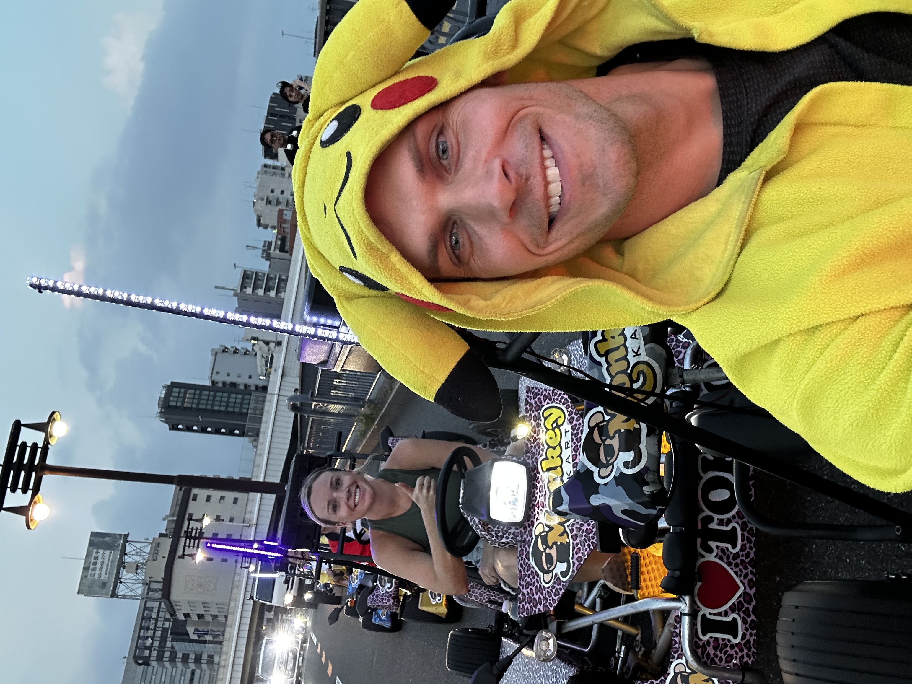 Driver in a Pikachu onesie in a purple camo go-kart beside another kart, with the Tokyo Skytree at dusk