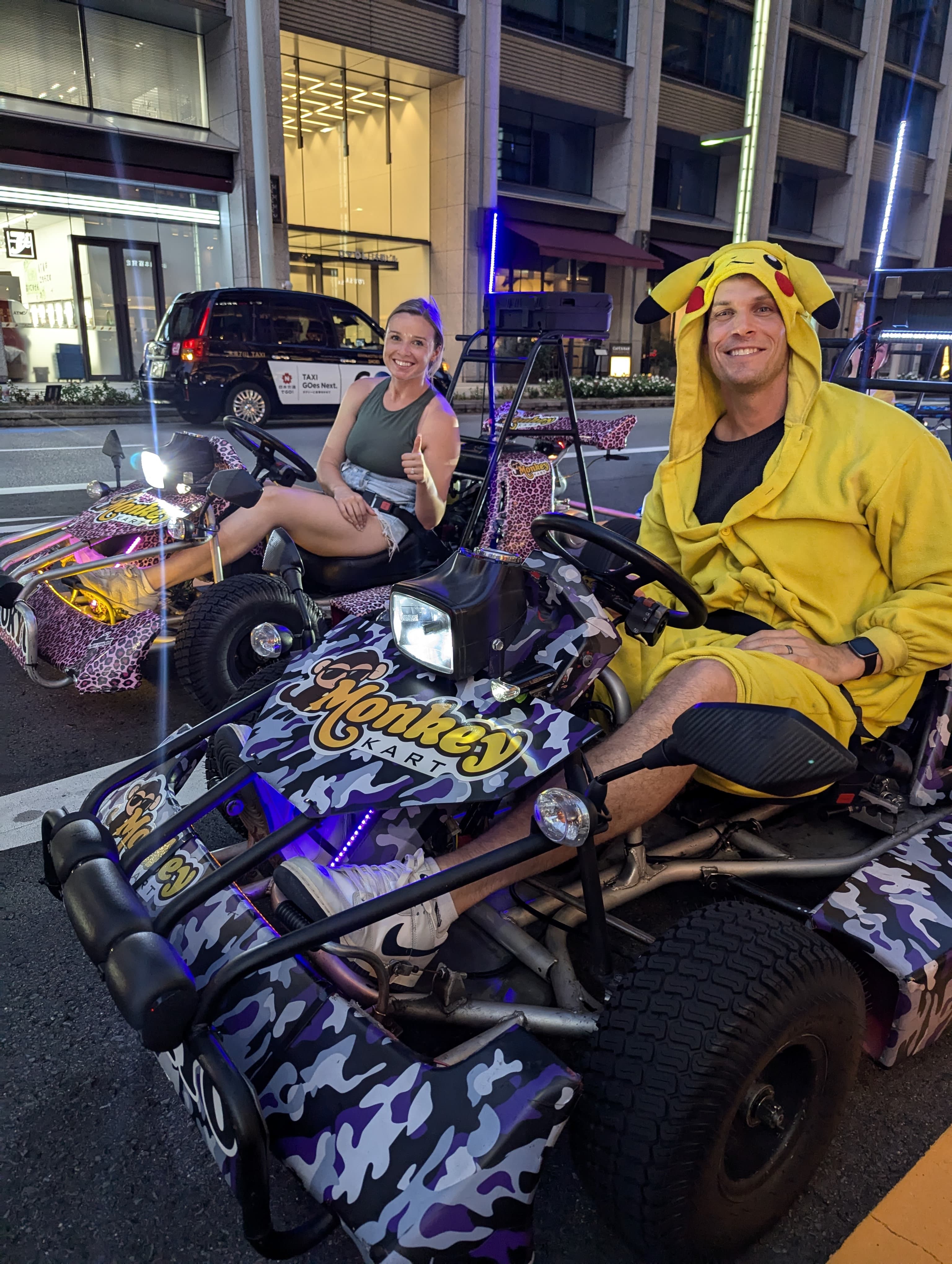 Two drivers in costume go-karts, one in a Pikachu onesie and one in a pink leopard-print kart, on a Tokyo street at twilight