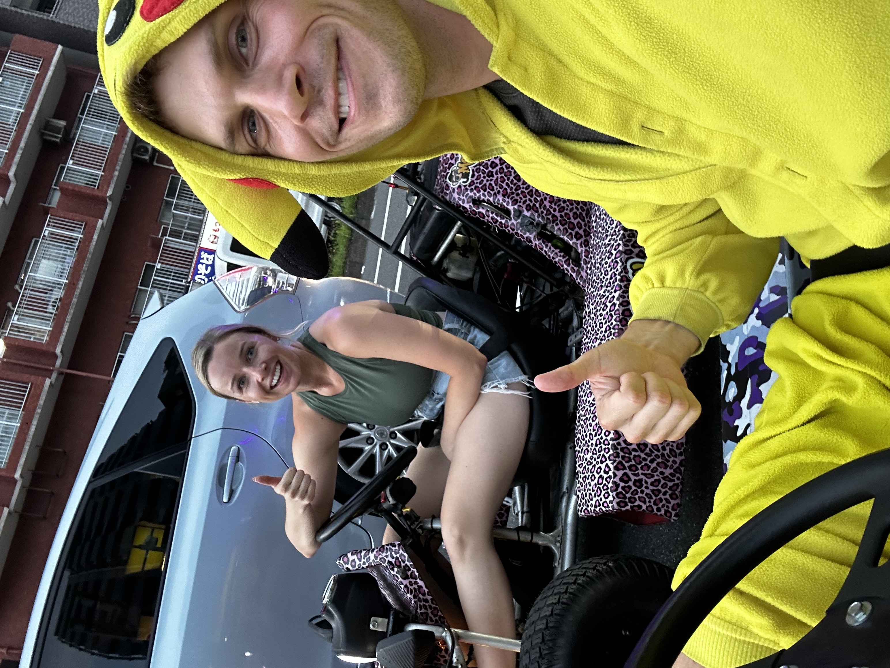 Two go-kart drivers giving a thumbs up from their karts, both smiling, parked on a Tokyo street
