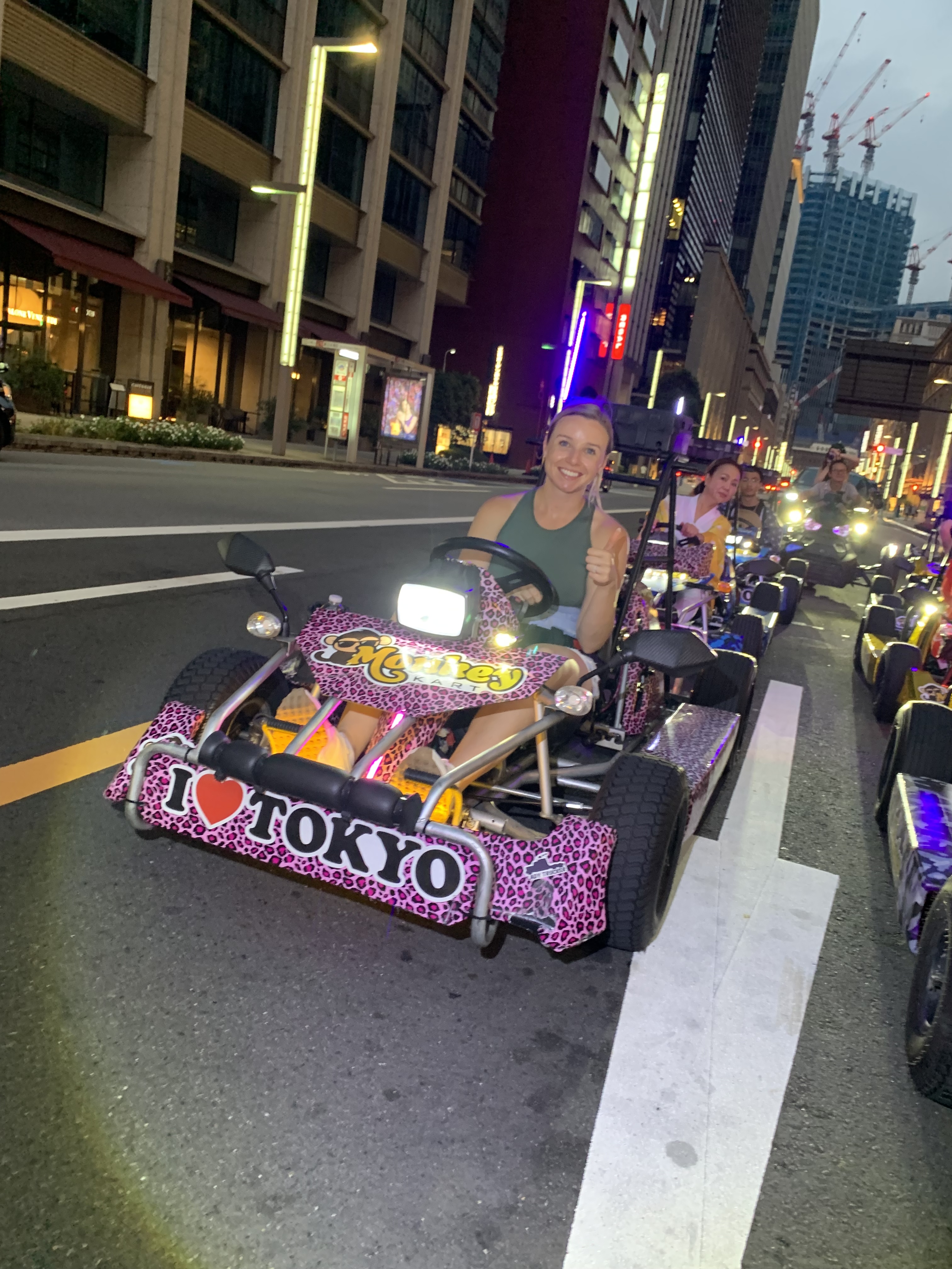 Driver in a pink leopard-print go-kart giving a thumbs up, with a convoy of go-karts on a Tokyo street at dusk