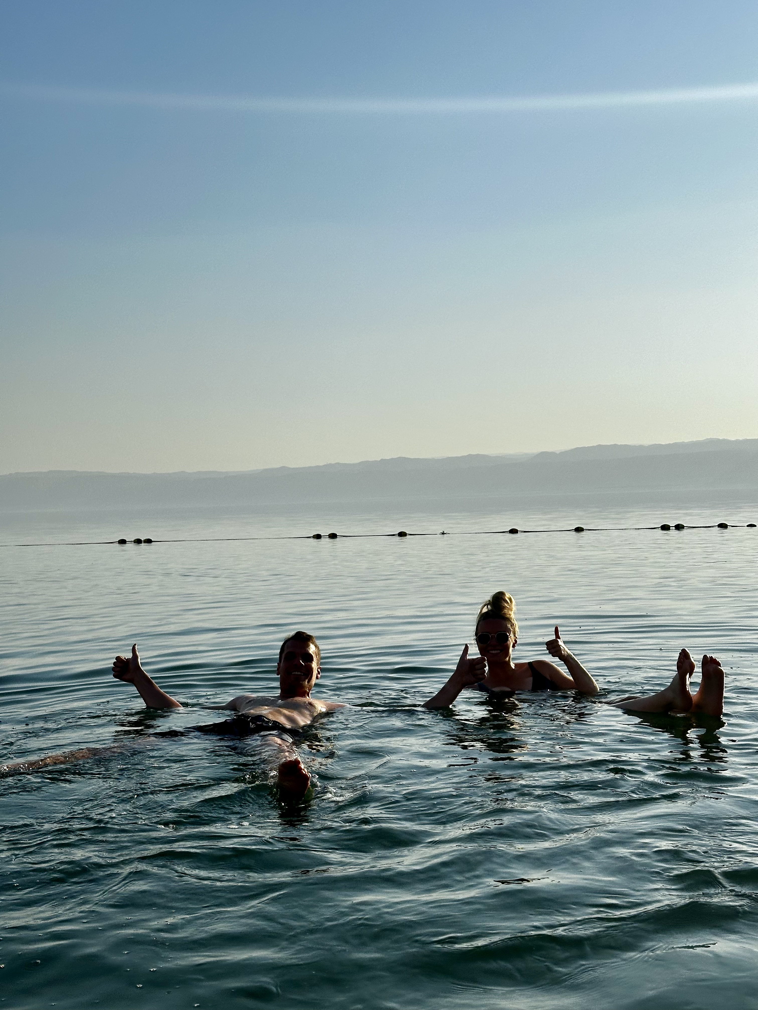 Two people floating on their backs in the Dead Sea, giving a thumbs up