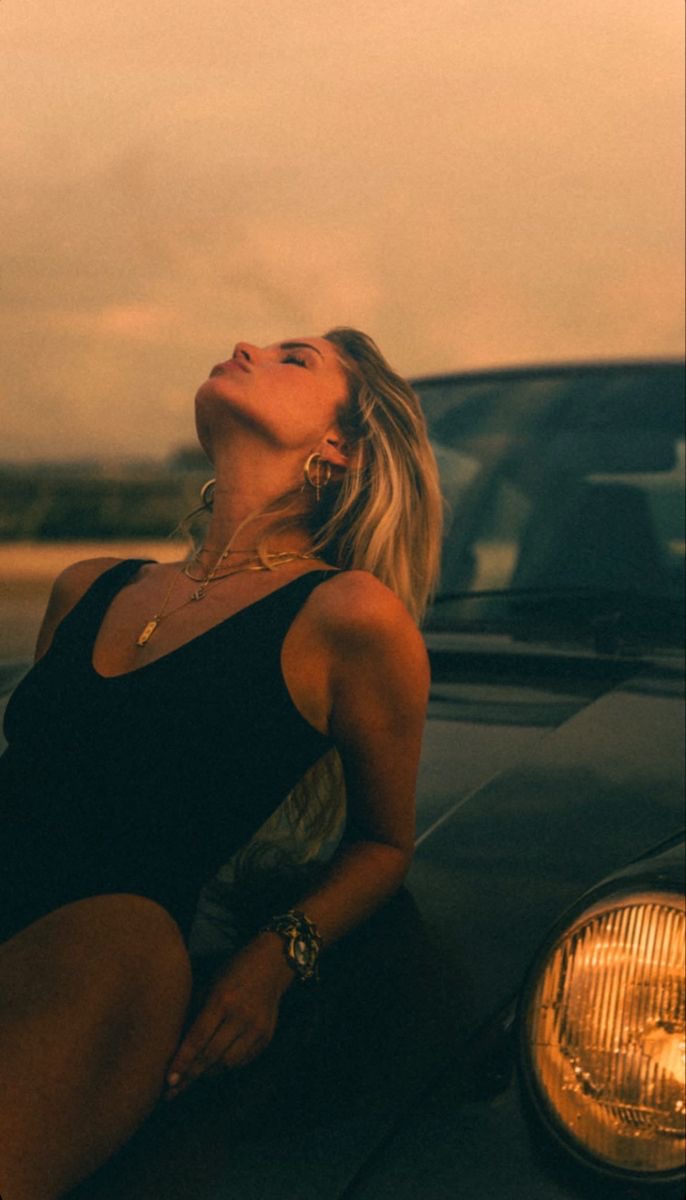 Woman leaning back on the hood of a vintage car at dusk