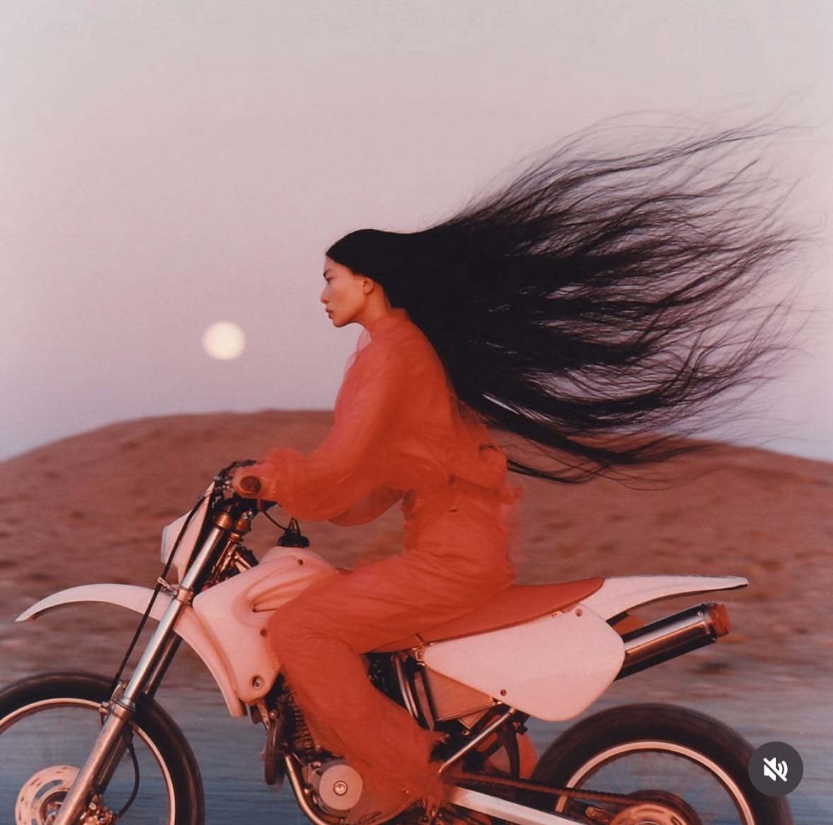 Woman with flowing dark hair riding a white dirt bike across a desert at dusk