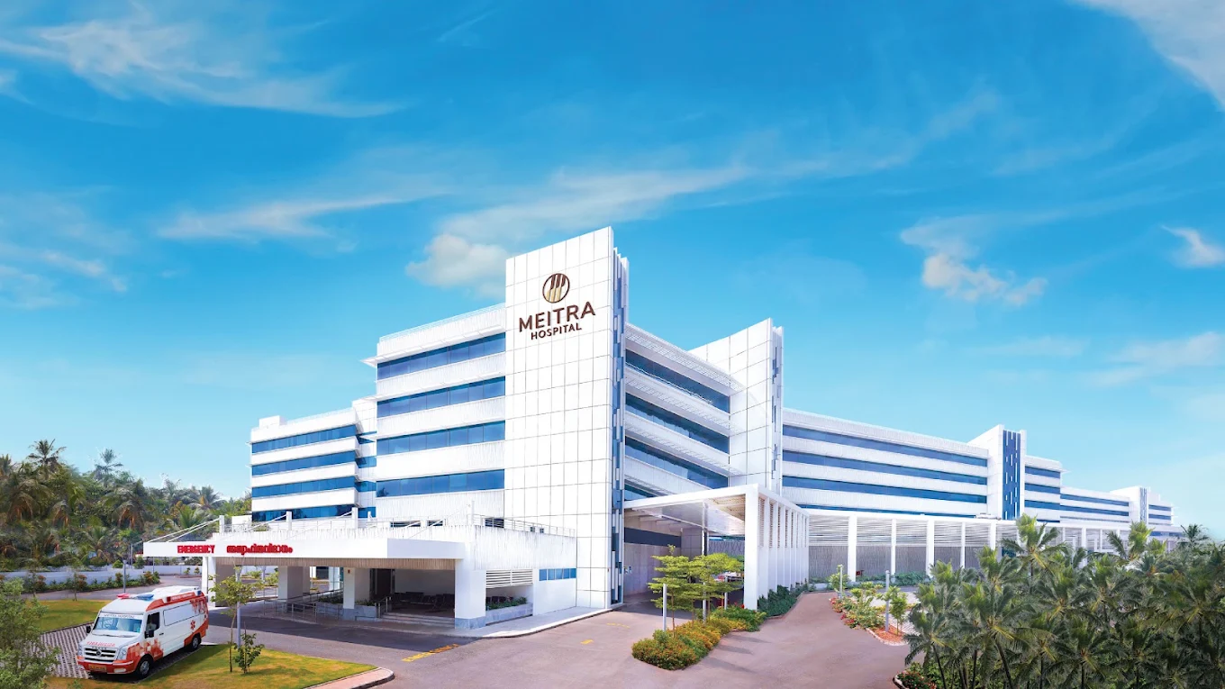 Meitra Hospital exterior architectural view showing the modern multi-story hospital building with white grid-paneled facade and dark reflective glass windows, set against a clear blue sky with landscaped surroundings.