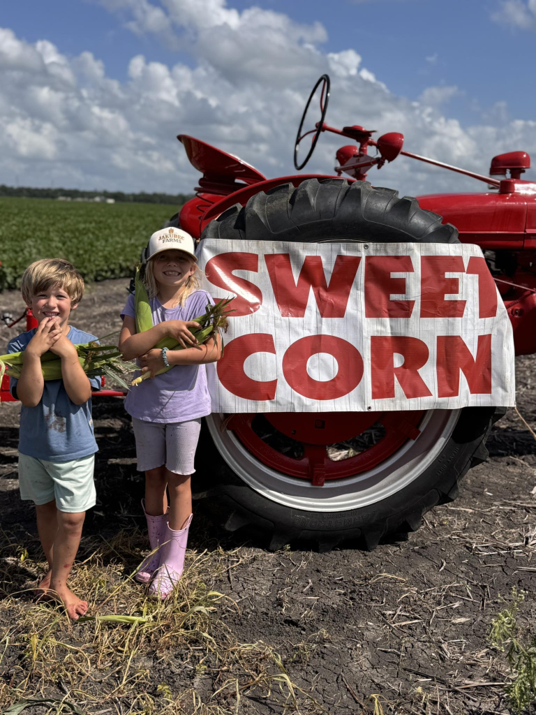 Kids holding sweet corn by the red tractor and sign