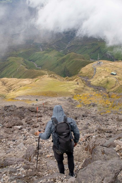 A hiker viewed from behind, wearing a slate-blue windbreaker and black backpack, descending a steep rocky mountain path marked with orange poles, overlooking a foggy valley of alpine tussock grass and green slopes below.