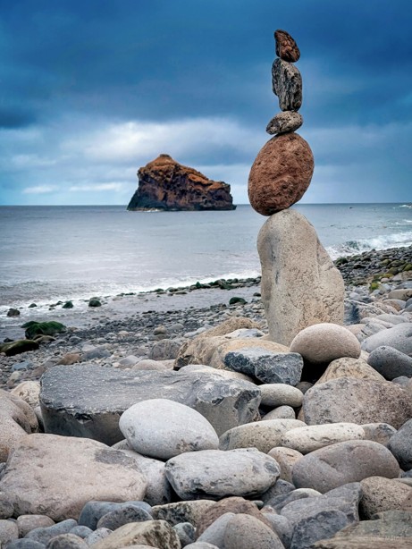 A stone balance sculpture stacked on a rocky beach, with a calm grey-blue ocean and an overcast sky behind it.