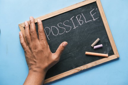 A top-down view of a small rectangular chalkboard with a light wood frame on a light-blue background, with the word POSSIBLE written in white chalk; a hand extends over the left edge transforming IMPOSSIBLE into POSSIBLE, with three coloured chalk pieces resting on the board.
