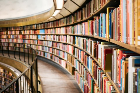 A high-angle long-shot of a curved library bookshelf spanning multiple levels, with sharply focused colourful books in the foreground on the right and shelves curving into a softly blurred distance on the left, warm wood tones and recessed track lighting above.