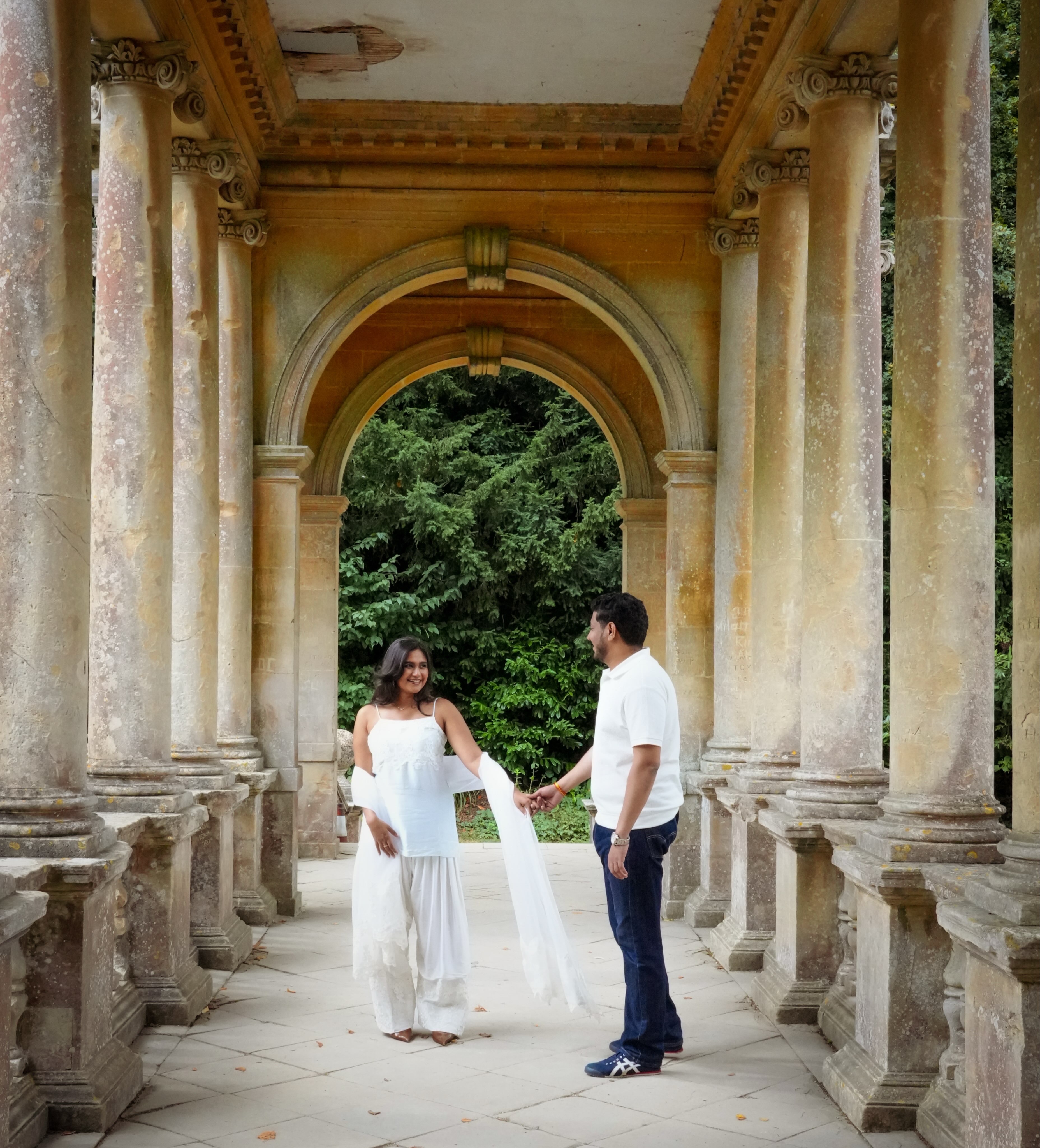 Couple holding hands in stone colonnade with green trees beyond