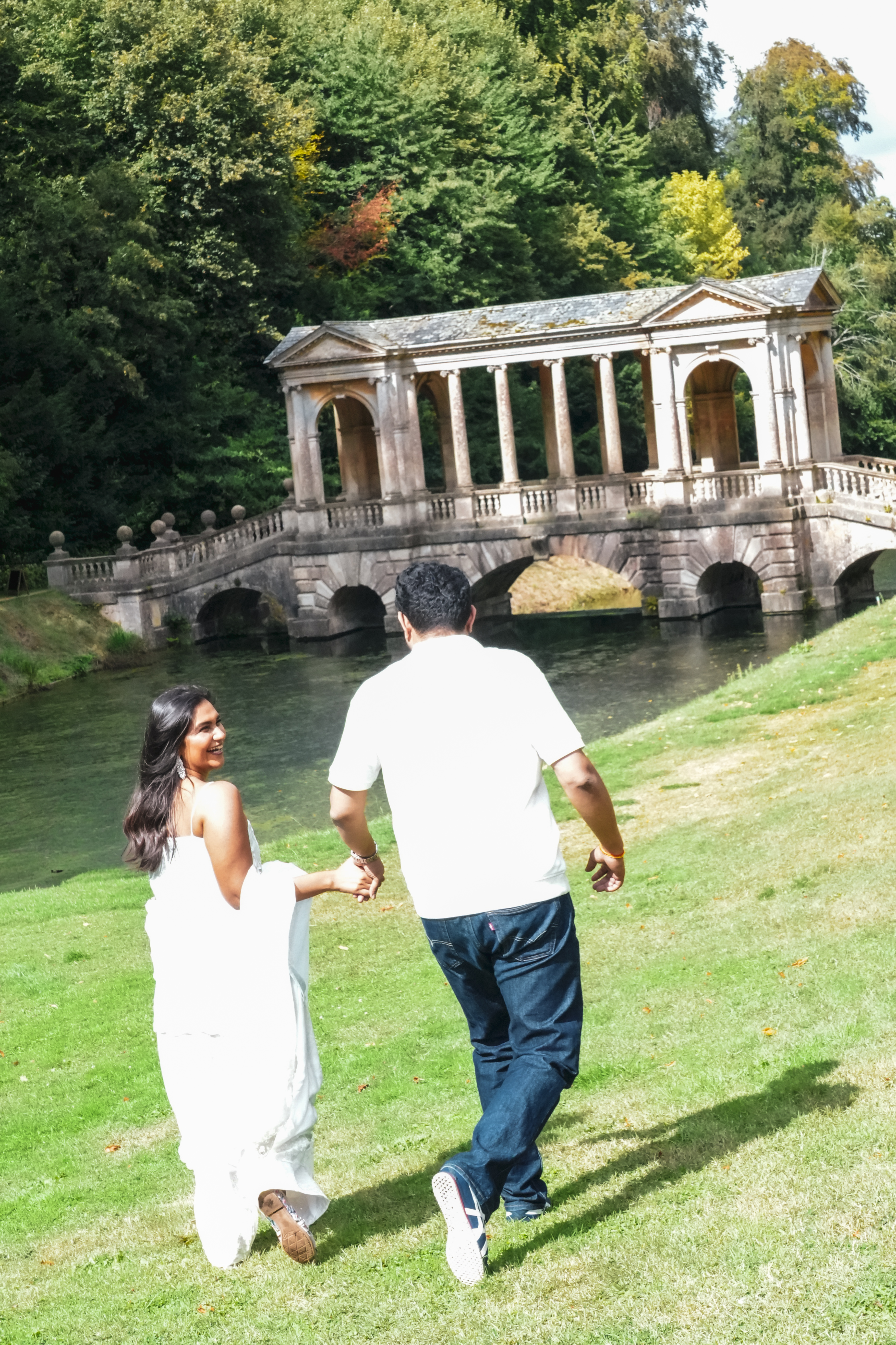 Couple walking hand in hand across grassy field toward Palladian bridge