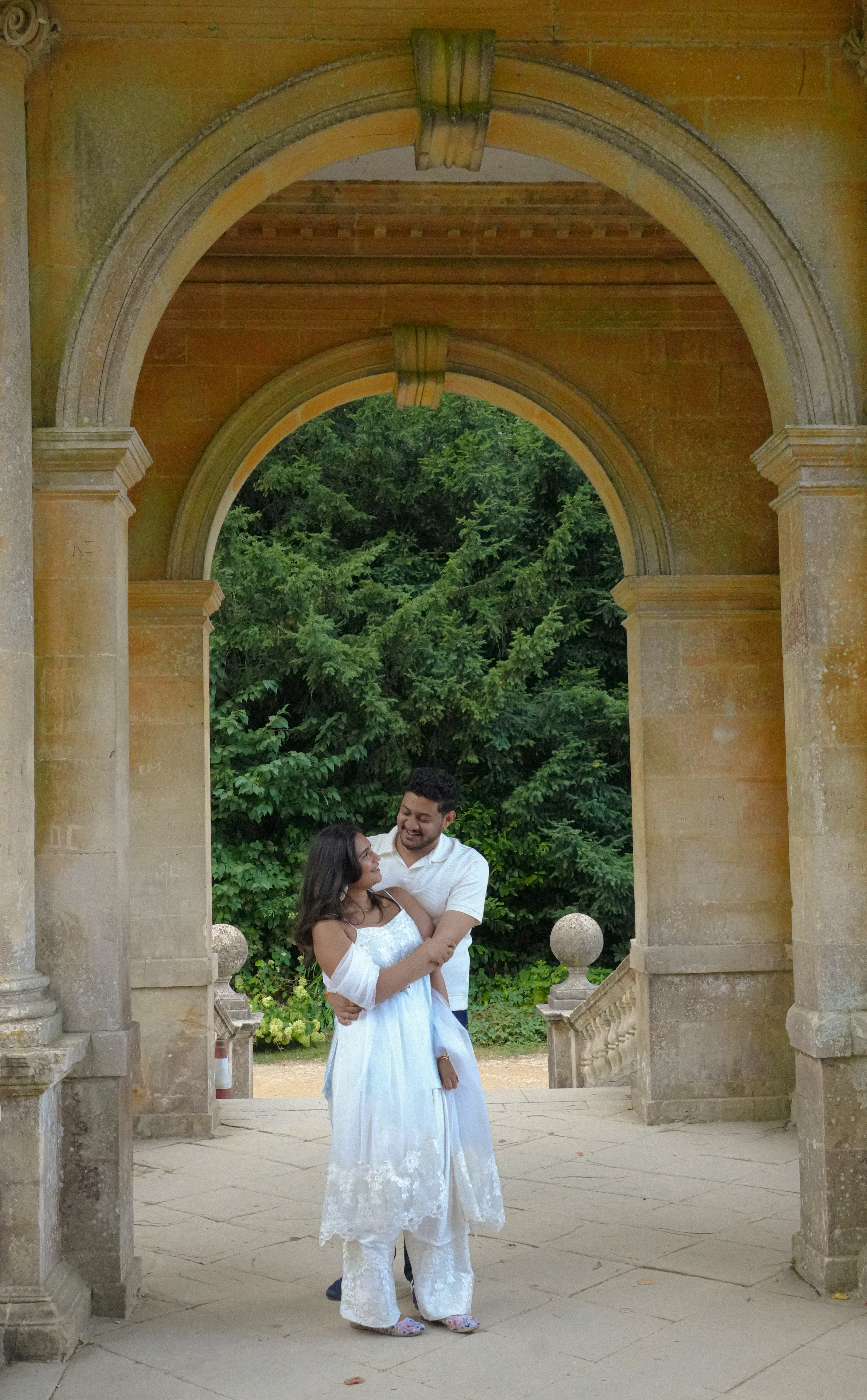 Couple embracing in stone archway with green trees behind