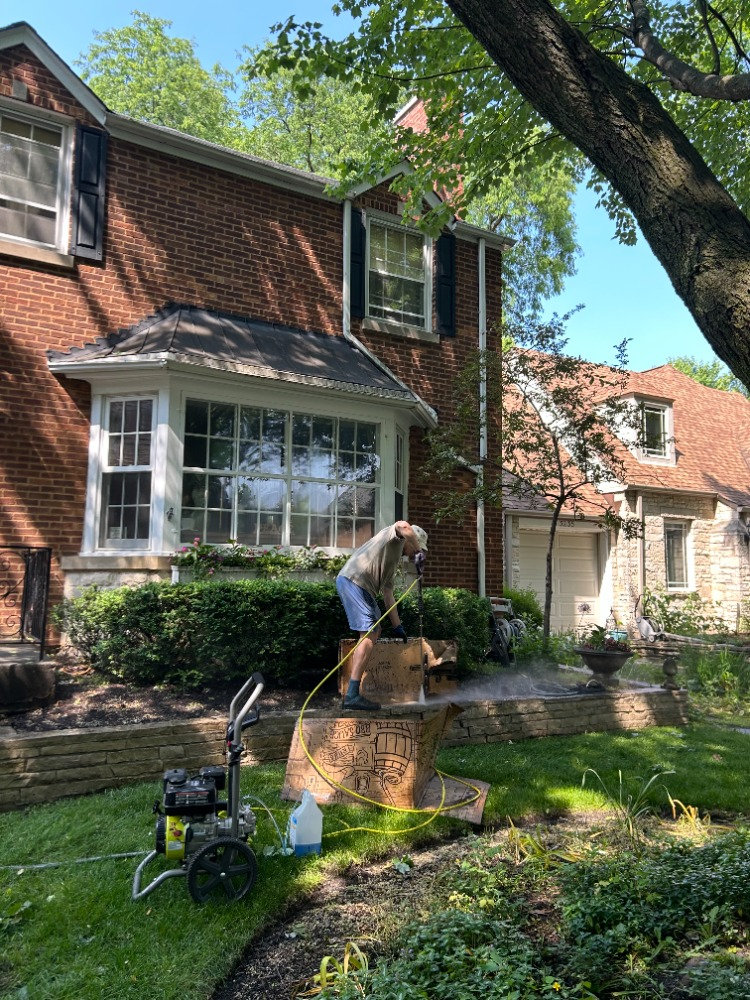 Teo power washing a planter box in front of a brick home