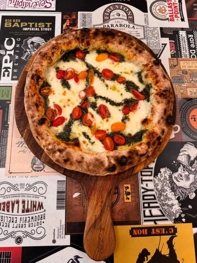 Pizza pronta com tomate e folhas de manjericão sobre uma pá de madeira, da Varanda 228.