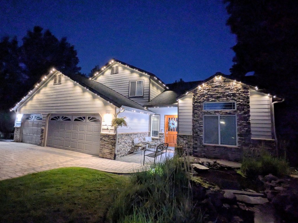 Two-story home with warm white Gemstone Lights architectural roofline lighting and stone facade at dusk in Central Oregon